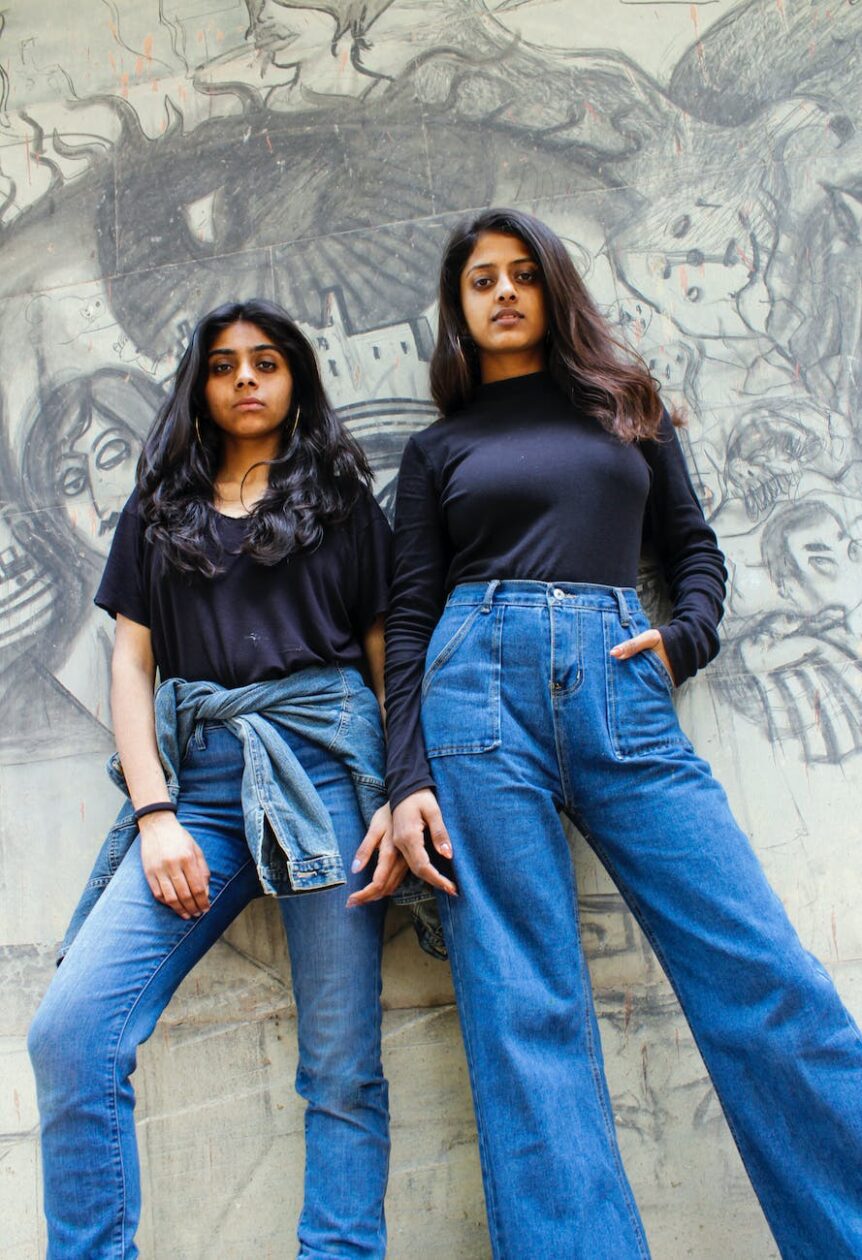 two women wearing black tops and blue jeans leaning on concrete wall